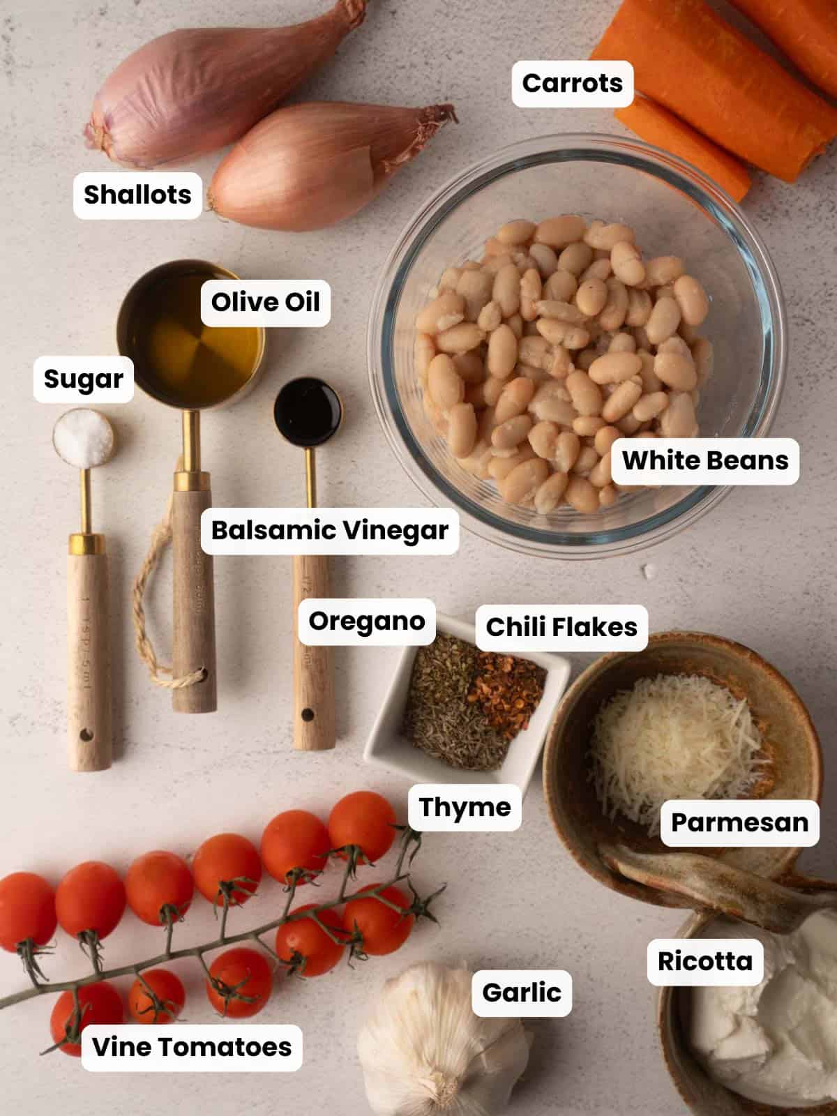 A flat lay photograph of fresh ingredients for the tomato and carrot soup recipe, featuring cannellini beans, shallots, carrots, cherry tomatoes, garlic, Parmesan cheese, and spices arranged on a light surface.