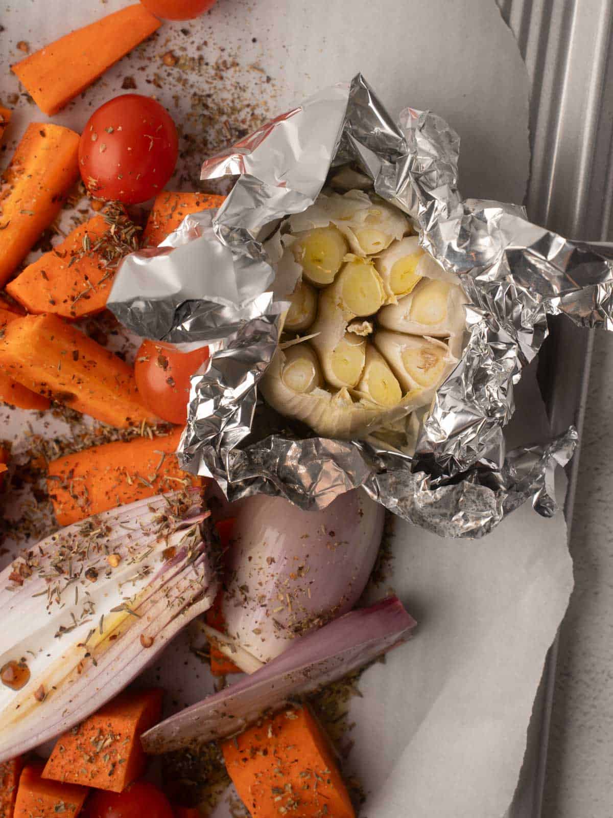 A close-up shot of seasoned vegetables on a parchment-lined baking sheet, featuring chopped carrots, cherry tomatoes, shallots, and a head of garlic wrapped in foil, ready for roasting.