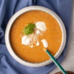 Overhead view of a bowl of creamy tomato carrot soup on a blue cloth, garnished with ricotta and Parmesan.