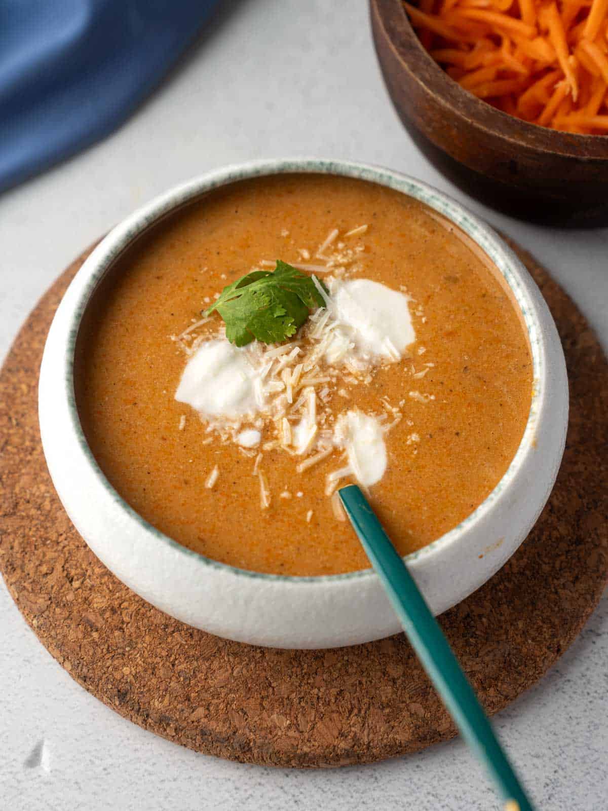 A close-up shot of a white bowl filled with creamy tomato carrot soup, garnished with dollops of sour cream, shredded Parmesan cheese, and a fresh cilantro leaf.