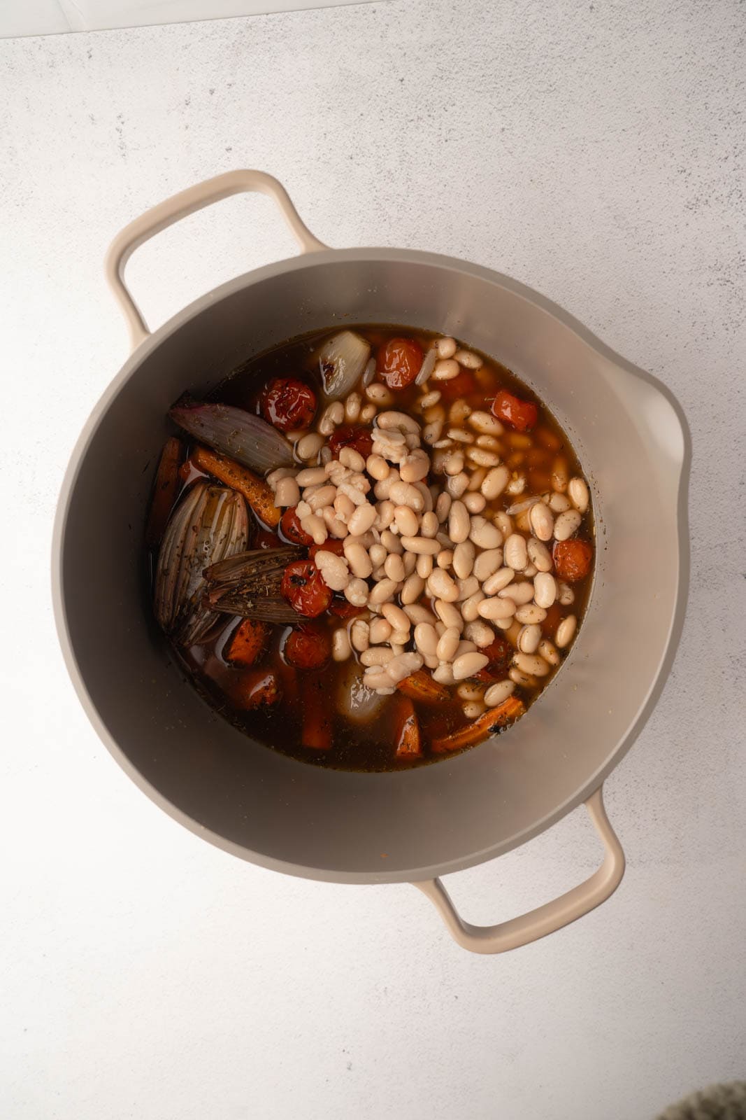 A top-down view of a soup pot filled with cooked cannellini beans, roasted vegetables, and broth, for tomato carrot soup preparation.