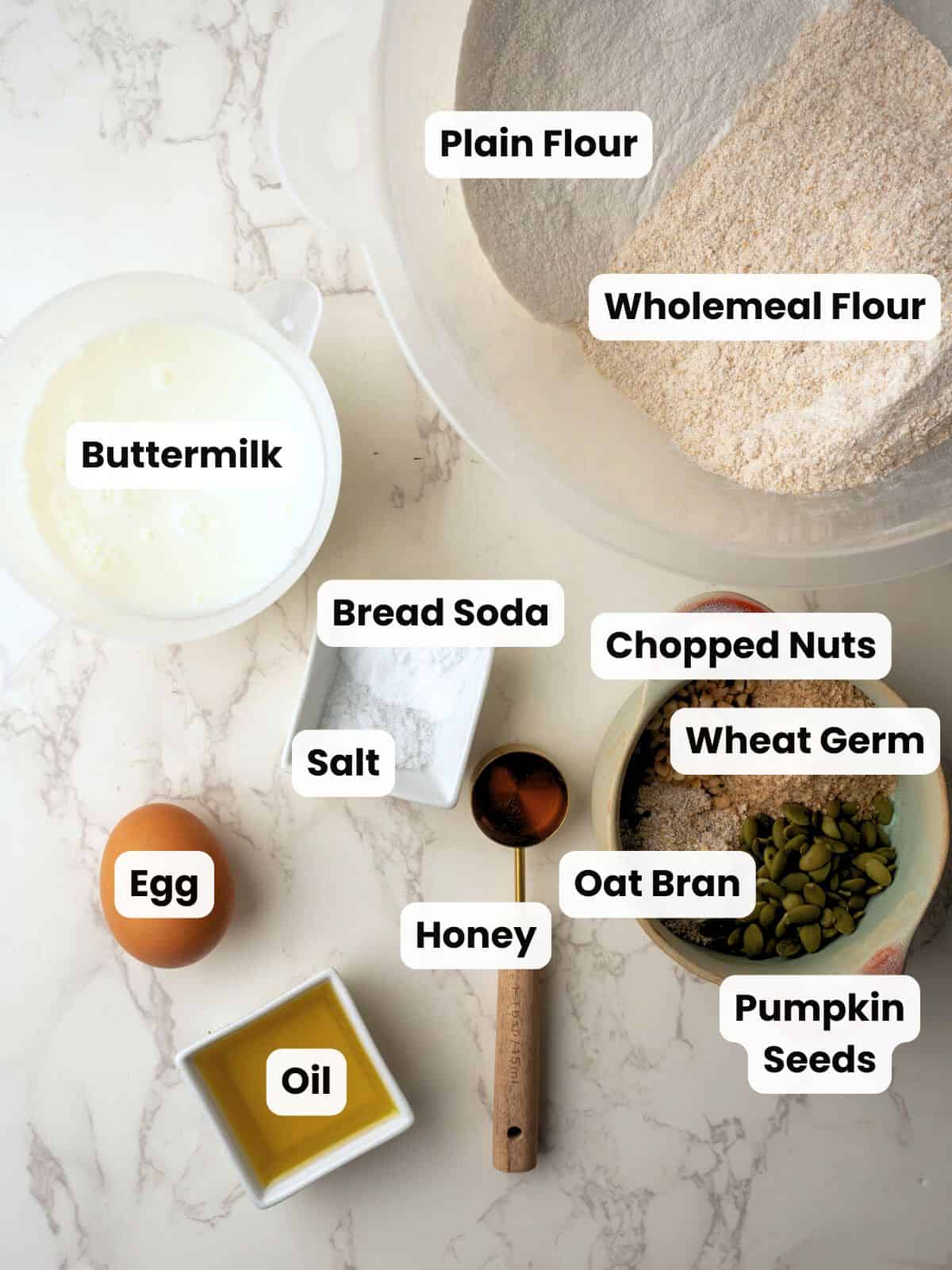 Overhead view of Irish brown bread ingredients on a marble surface, including plain flour, wholemeal flour, buttermilk, bread soda, salt, egg, oil, honey, oat bran, wheat germ, pumpkin seeds, and chopped nuts, each labeled with white text boxes.