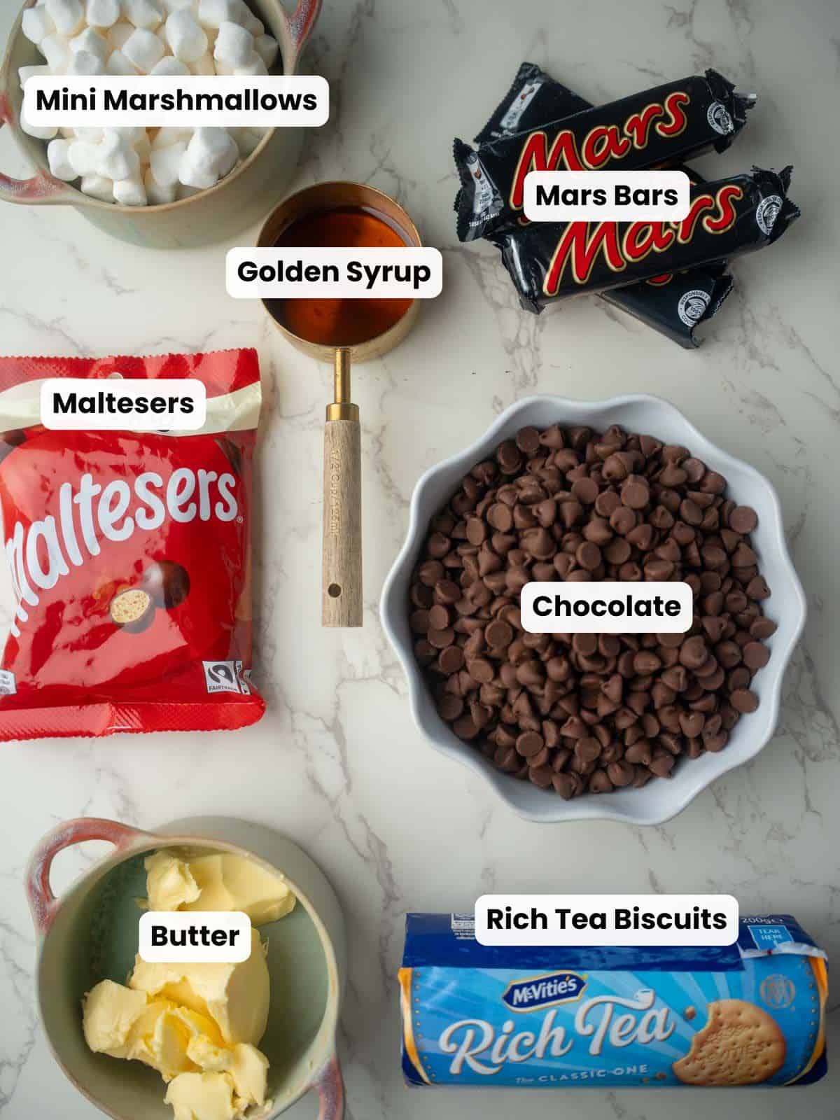 Flatlay of ingredients used to make rocky road with Maltesers, including rich tea biscuits, mini marshmallows, chocolate chips, butter, Maltesers, Mars bars, and golden syrup.