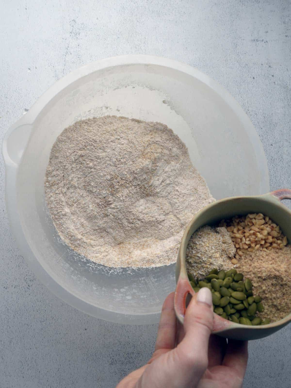 Hand holding a small bowl of pumpkin seeds, chopped nuts, oat bran, and wheat germ over a large mixing bowl filled with flour.