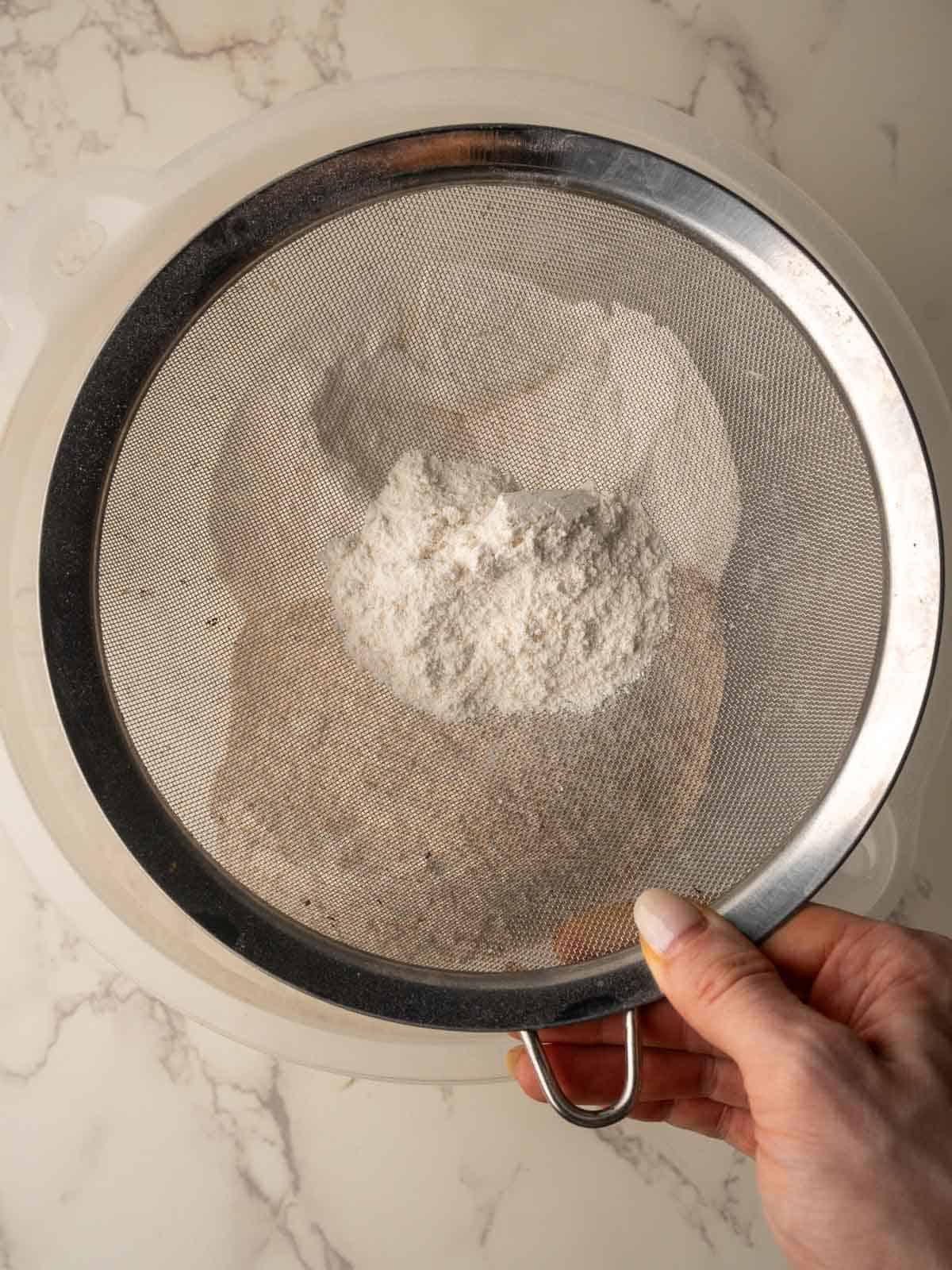 Hand holding a sieve over a mixing bowl, sifting flour for Irish brown bread on a marble countertop.