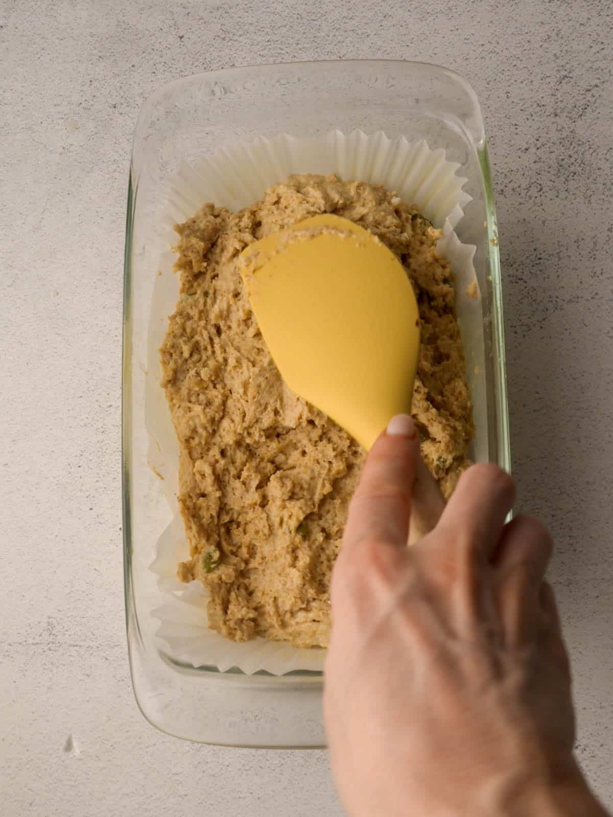 Hand using a yellow spatula to spread Irish brown bread dough evenly into a parchment-lined glass loaf pan.