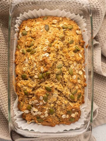 Close-up overhead view of a freshly baked Irish brown bread loaf in a glass pan lined with parchment paper, topped with oats and pumpkin seeds, resting on a beige kitchen towel.