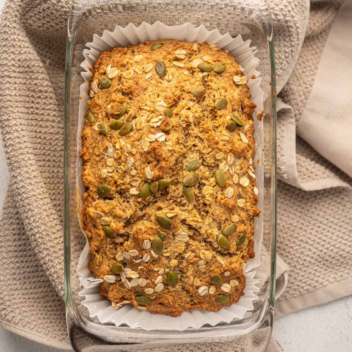 Close-up overhead view of a freshly baked Irish brown bread loaf in a glass pan lined with parchment paper, topped with oats and pumpkin seeds, resting on a beige kitchen towel.