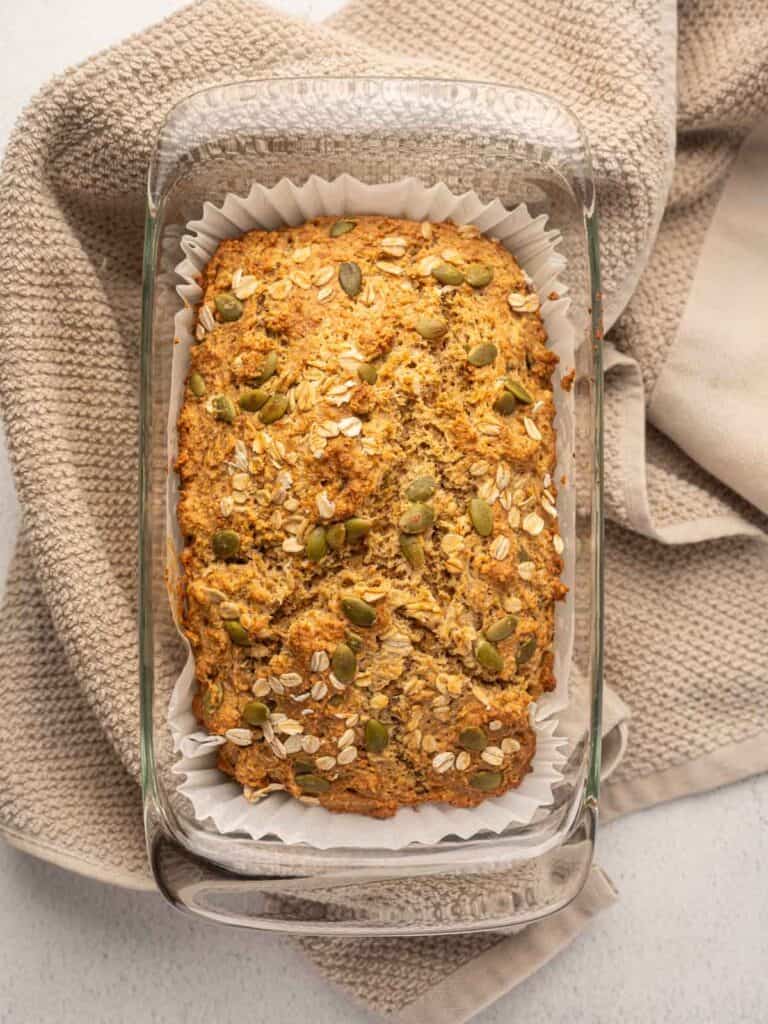 Close-up overhead view of a freshly baked Irish brown bread loaf in a glass pan lined with parchment paper, topped with oats and pumpkin seeds, resting on a beige kitchen towel.