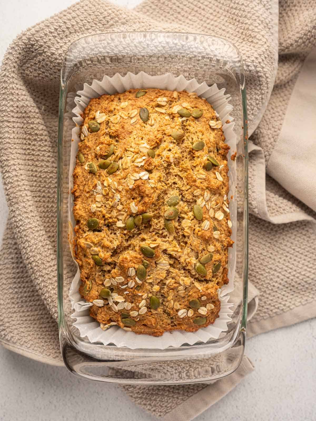 Freshly baked Irish brown bread in a glass loaf pan lined with parchment paper, topped with oats and pumpkin seeds, resting on a beige textured kitchen towel.