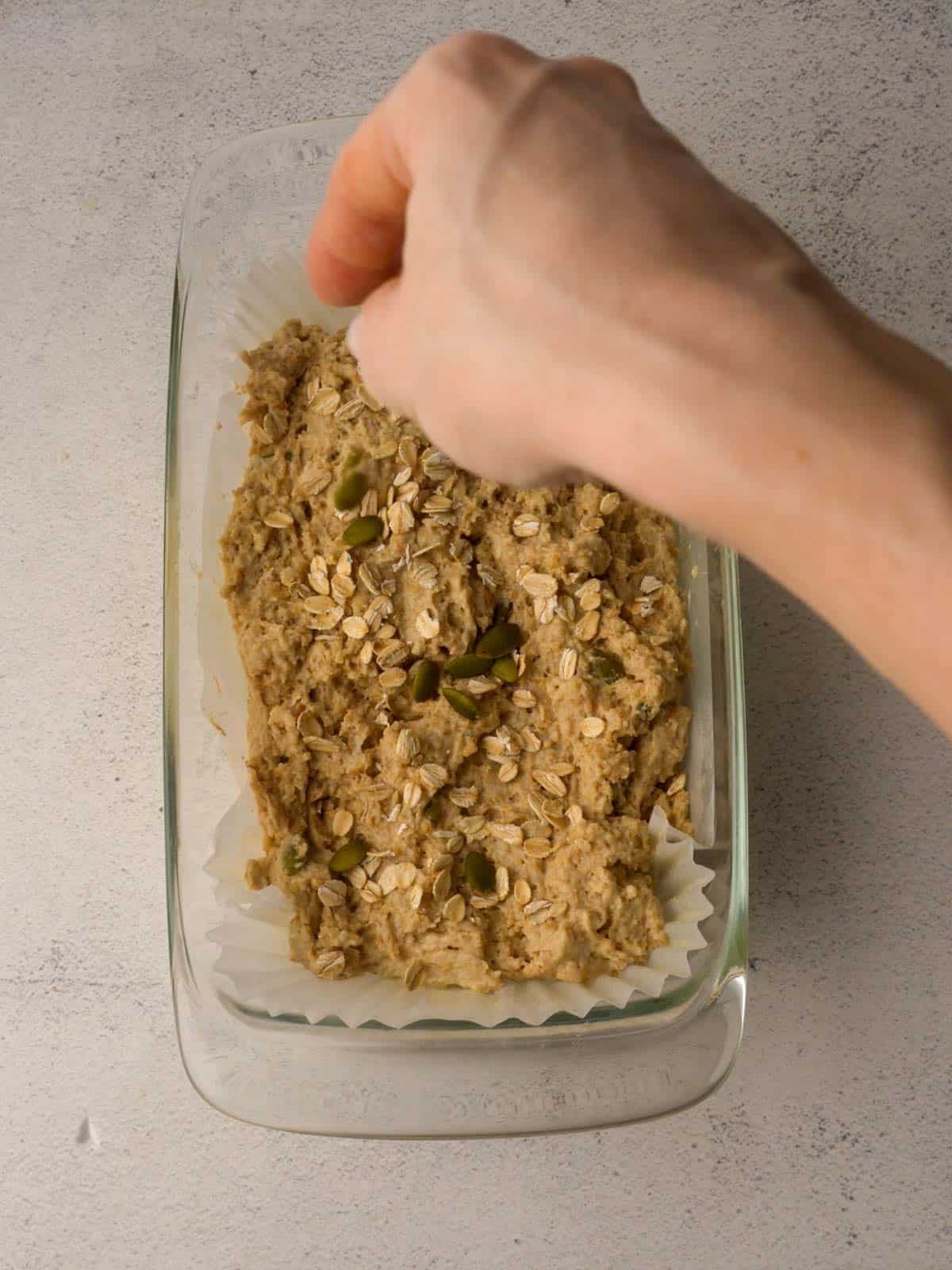 Hand sprinkling oats and pumpkin seeds over unbaked brown bread batter in a glass loaf pan.