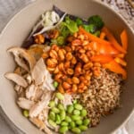 Bowl of spicy chicken quinoa salad with shredded chicken, edamame, bell pepper, slaw mix, cilantro, quinoa, and honey-sriracha peanuts arranged in sections, shot overhead on a neutral background.