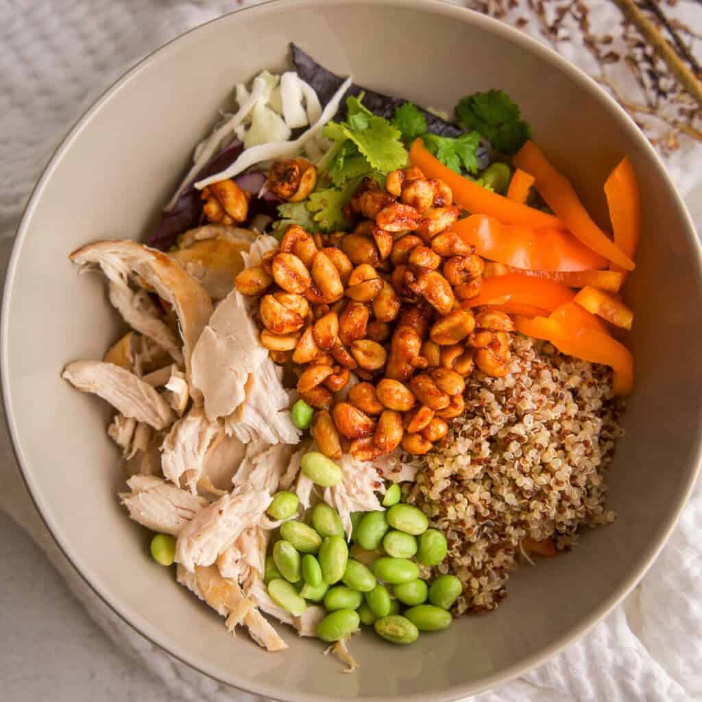 Bowl of spicy chicken quinoa salad with shredded chicken, edamame, bell pepper, slaw mix, cilantro, quinoa, and honey-sriracha peanuts arranged in sections, shot overhead on a neutral background.