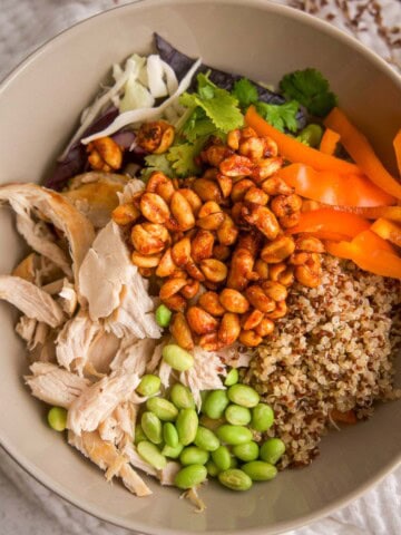 Bowl of spicy chicken quinoa salad with shredded chicken, edamame, bell pepper, slaw mix, cilantro, quinoa, and honey-sriracha peanuts arranged in sections, shot overhead on a neutral background.