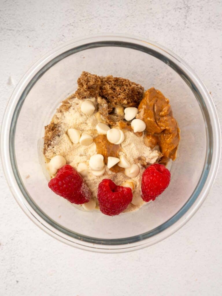 Glass mixing bowl filled with Weetabix, peanut butter, protein powder, white chocolate chips, and fresh raspberries before mixing, set on a light countertop.