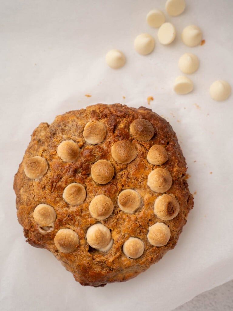 Overhead view of a golden Weetabix cake topped with toasted white chocolate chips, placed on parchment paper with a few white chocolate chips scattered nearby.