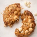 Close-up of a golden-brown baked Weetabix cake topped with lightly toasted white chocolate chips, resting on parchment paper , broken in half, with a few extra chocolate chips scattered around.