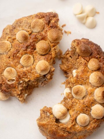 Close-up of a golden-brown baked Weetabix cake topped with lightly toasted white chocolate chips, resting on parchment paper , broken in half, with a few extra chocolate chips scattered around.