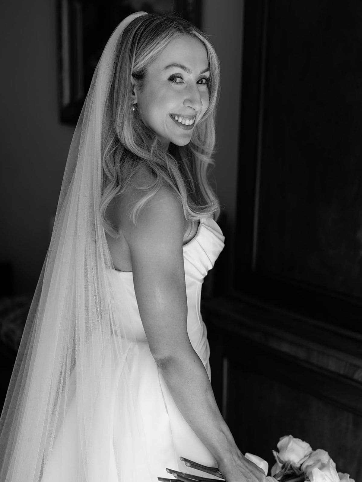 Smiling bride in a strapless wedding dress and long veil, looking over her shoulder while holding a bouquet, black and white photo.