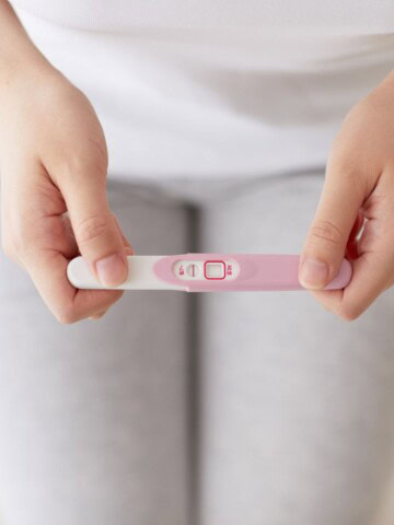 Person holding a home pregnancy test with one visible line, seated with gray leggings in the background.