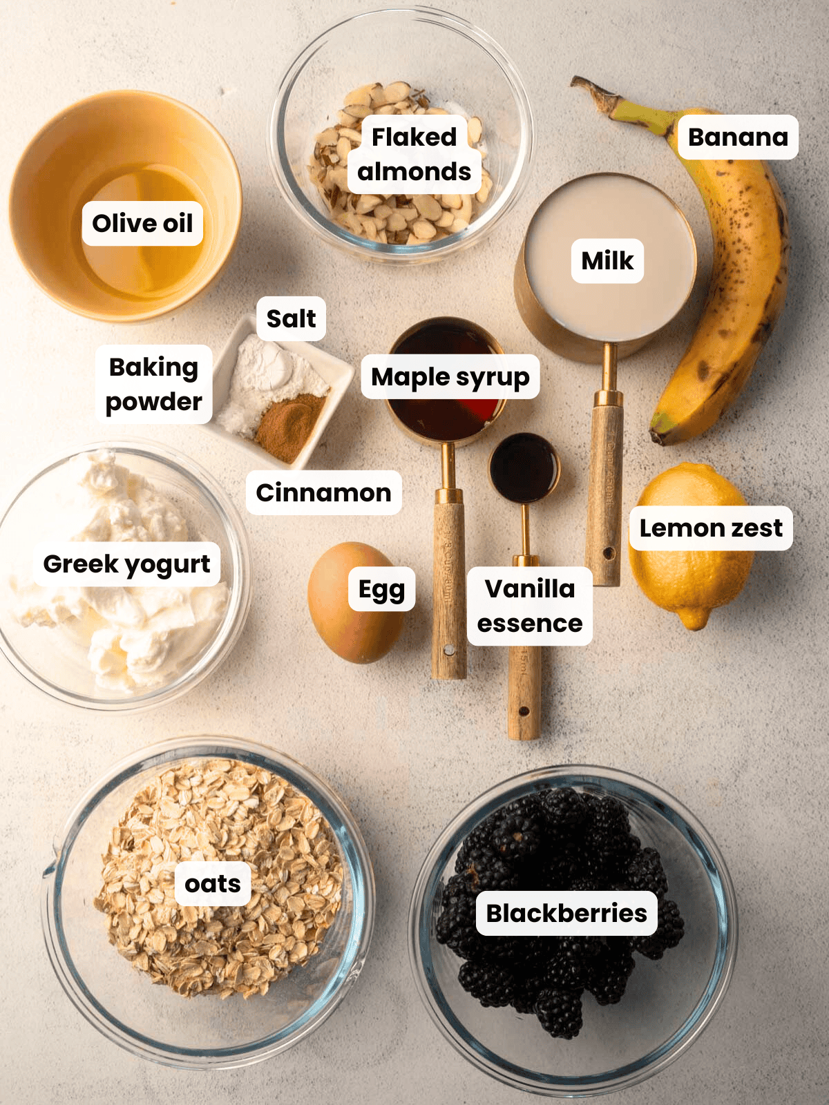 Flat-lay of labeled ingredients for blackberry baked oatmeal, including oats, blackberries, Greek yogurt, banana, milk, flaked almonds, egg, olive oil, maple syrup, vanilla essence, lemon zest, cinnamon, baking powder, and salt.