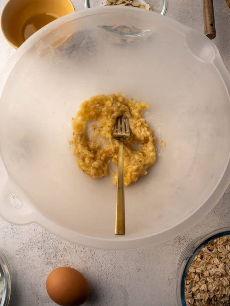Mashed banana in a large mixing bowl with a gold fork, surrounded by ingredients like oats, eggs, and almonds ready to be added.