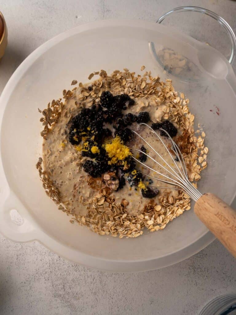 Oatmeal mixture in a mixing bowl with blackberries, oats, and lemon zest being stirred with a whisk.