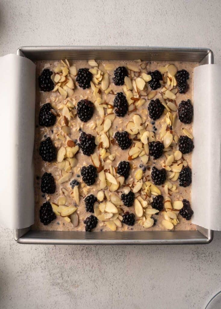 Unbaked blackberry oatmeal mixture in a square tin, topped with fresh blackberries and sliced almonds, ready for the oven.