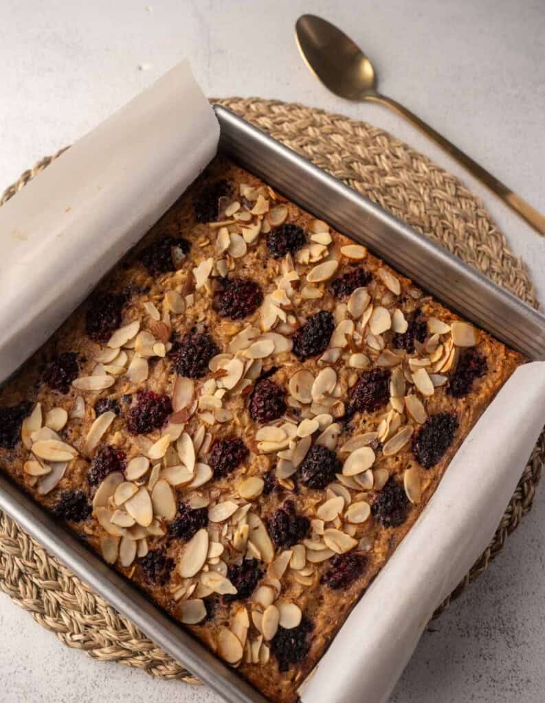 Baked blackberry oatmeal topped with toasted sliced almonds in a square tin lined with parchment, placed on a woven placemat with a gold spoon beside it.