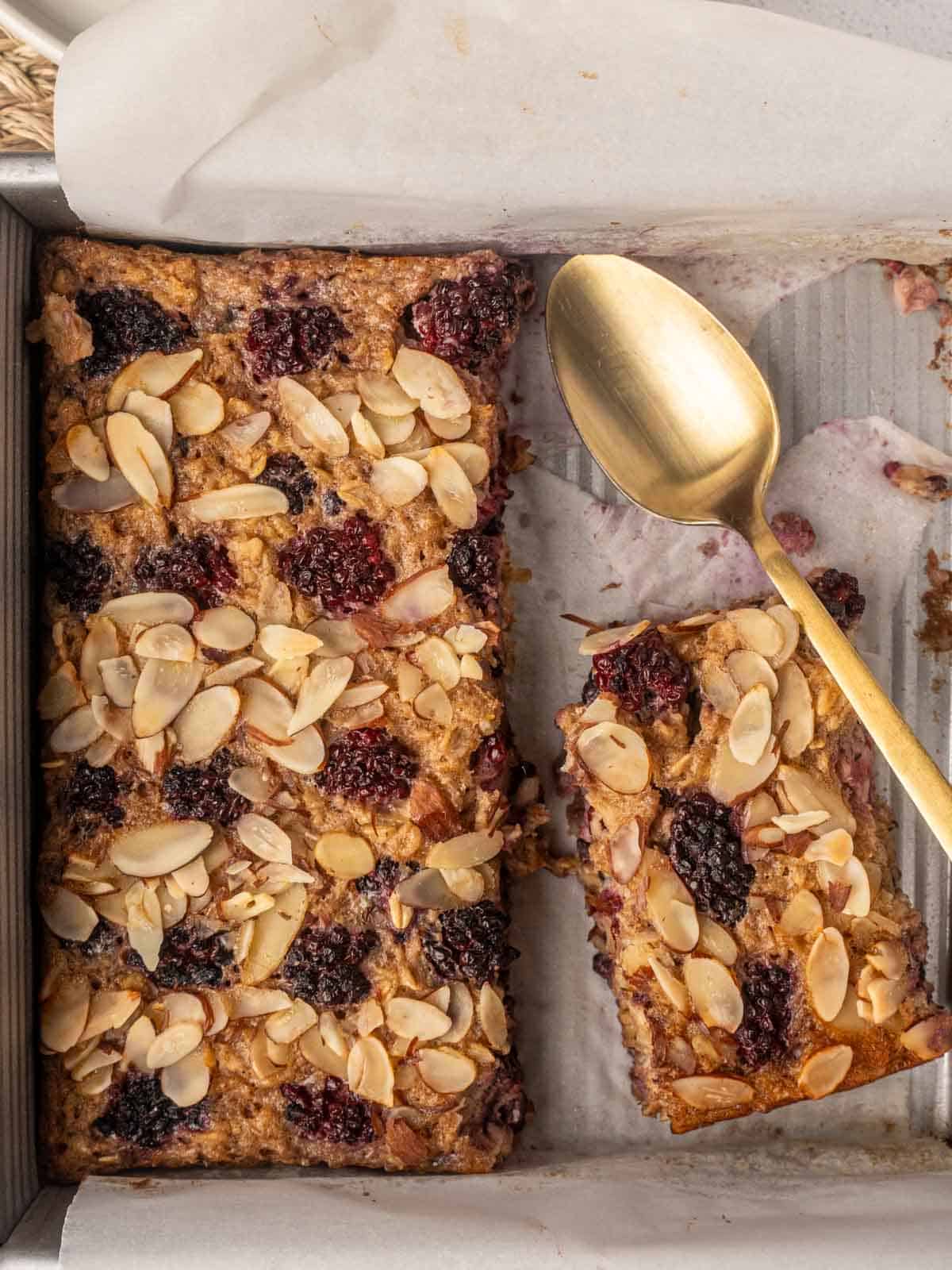 A partially sliced tray of blackberry baked oatmeal topped with toasted almond flakes, with a gold spoon resting on the parchment-lined baking tin.