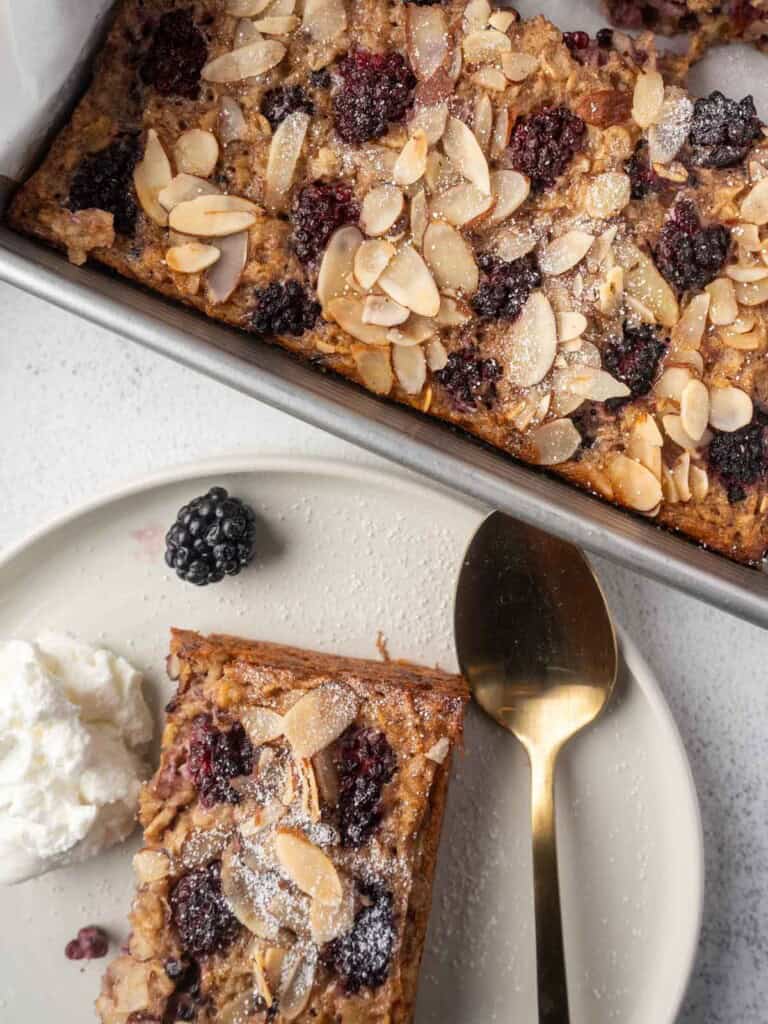 Blackberry baked oatmeal in a square baking dish topped with sliced almonds and powdered sugar, with a sliced piece served on a plate beside whipped yogurt and a fresh blackberry.
