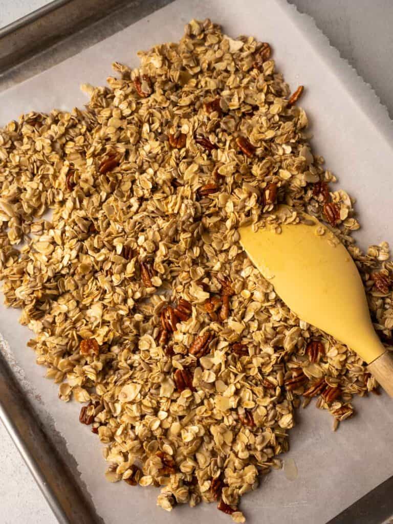 Unbaked granola mixture spread on a parchment-lined baking tray with a yellow spatula.