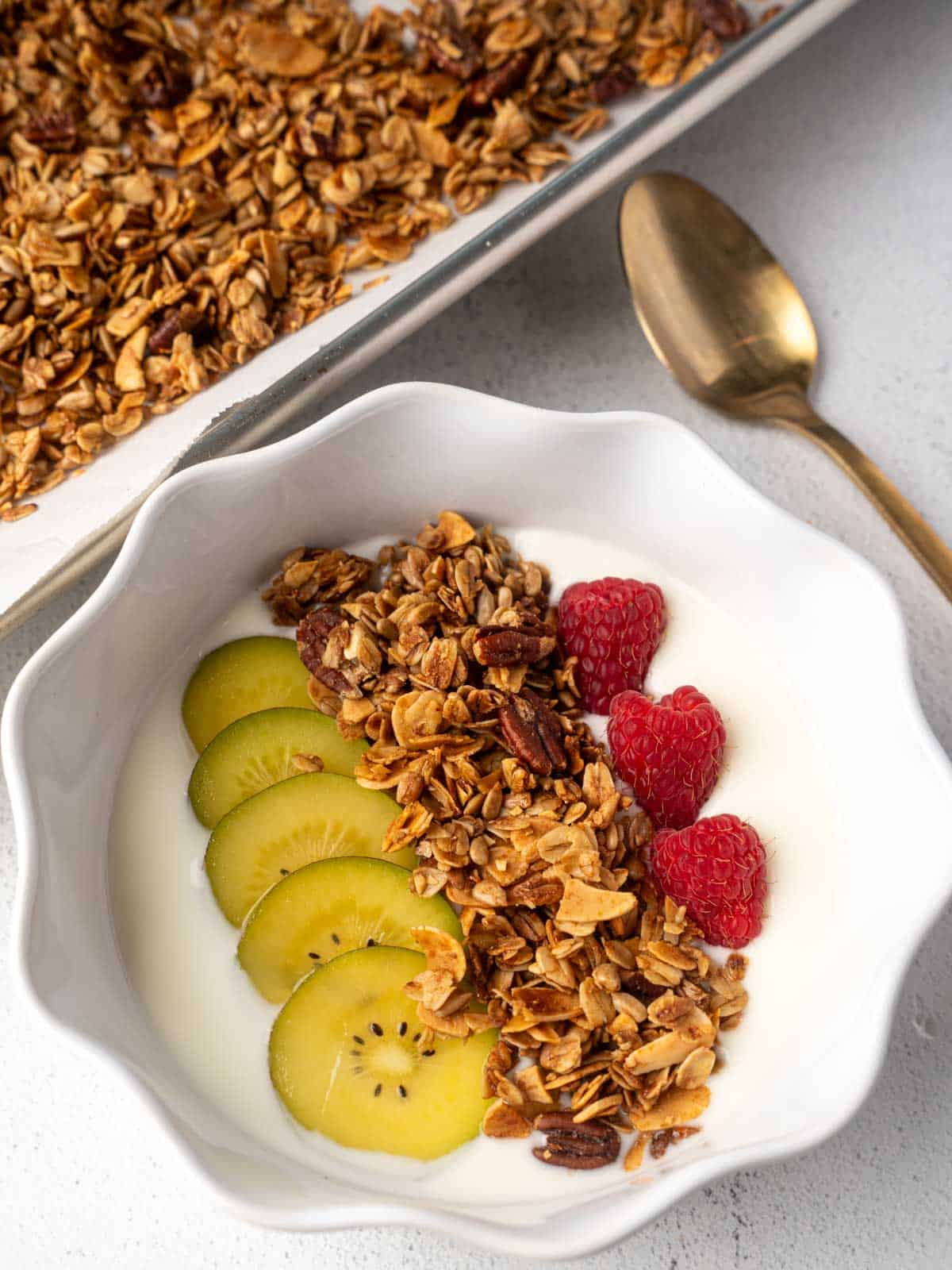 Bowl of yogurt topped with homemade granola, sliced golden kiwi, and fresh raspberries, with a tray of granola and a gold spoon in the background.