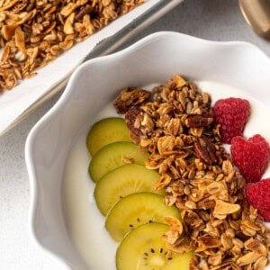 Bowl of yogurt topped with homemade granola, sliced golden kiwi, and fresh raspberries, with a tray of granola and a gold spoon in the background.
