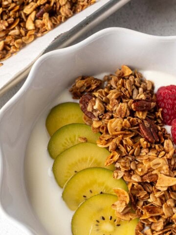 Bowl of yogurt topped with homemade granola, sliced golden kiwi, and fresh raspberries, with a tray of granola and a gold spoon in the background.
