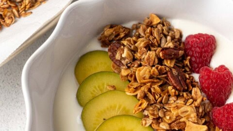 Bowl of yogurt topped with homemade granola, sliced golden kiwi, and fresh raspberries, with a tray of granola and a gold spoon in the background.