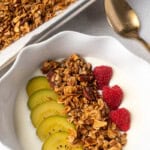 Bowl of yogurt topped with homemade granola, sliced golden kiwi, and fresh raspberries, with a tray of granola and a gold spoon in the background.