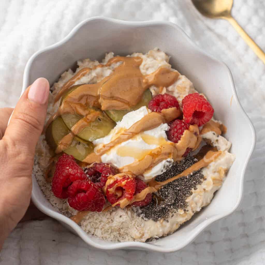 A white bowl filled with creamy high protein oatmeal topped with raspberries, sliced kiwi, chia seeds, a dollop of yogurt, and a drizzle of peanut butter and honey. A hand holds the bowl against a soft white background.