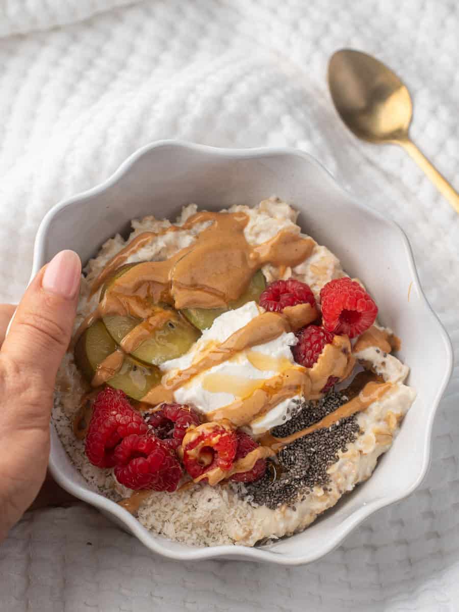 A white bowl filled with creamy high protein oatmeal topped with raspberries, sliced kiwi, chia seeds, a dollop of yogurt, and a drizzle of peanut butter and honey. A hand holds the bowl against a soft white background.