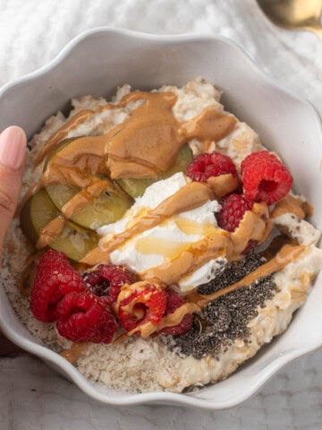A white bowl filled with creamy high protein oatmeal topped with raspberries, sliced kiwi, chia seeds, a dollop of yogurt, and a drizzle of peanut butter and honey. A hand holds the bowl against a soft white background.
