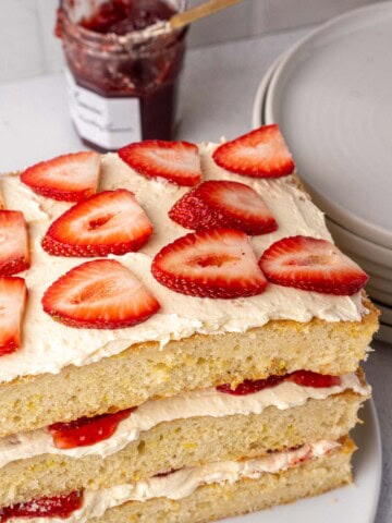 Strawberry layer cake with cream filling and jam, topped with fresh strawberries, served on a white cake stand with plates in the background.