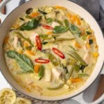 Thai green chicken curry in a white pan with coconut milk, bell peppers, zucchini, green beans, basil, and sliced red chili.
