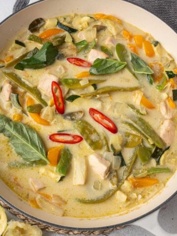 Thai green chicken curry in a white pan with coconut milk, bell peppers, zucchini, green beans, basil, and sliced red chili.