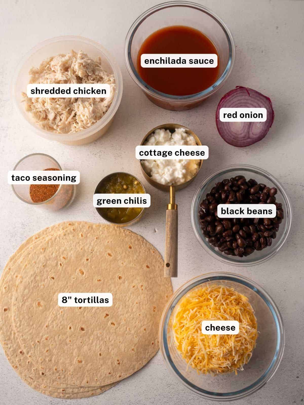 Ingredients for black bean chicken enchiladas arranged on a countertop including shredded chicken, enchilada sauce, red onion, taco seasoning, green chilis, cottage cheese, black beans, tortillas, and shredded cheese