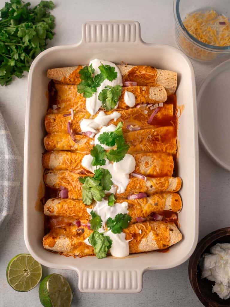 High-protein black bean chicken enchiladas baked in a white casserole dish, topped with melted cheese, sour cream, fresh cilantro, and diced red onion in a rich red enchilada sauce.