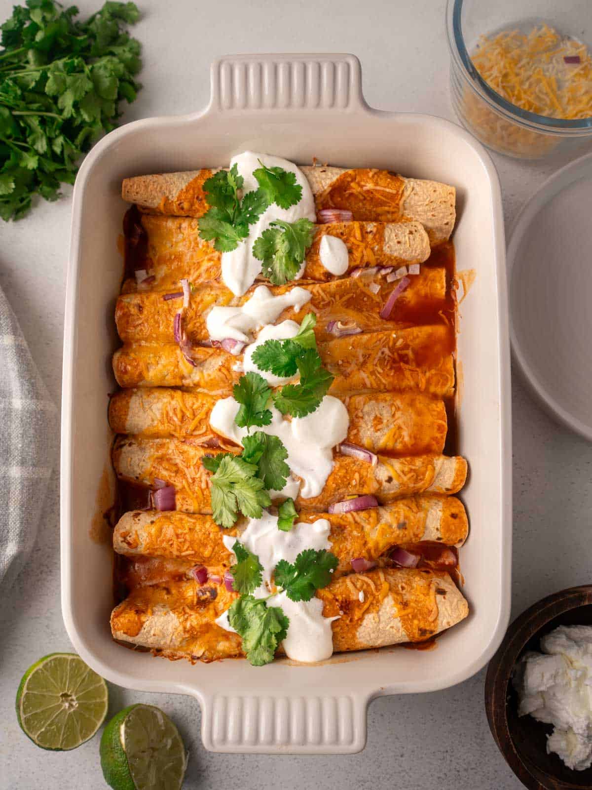 High-protein black bean chicken enchiladas baked in a white casserole dish, topped with melted cheese, sour cream, fresh cilantro, and diced red onion in a rich red enchilada sauce.