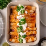High-protein black bean chicken enchiladas baked in a white casserole dish, topped with melted cheese, sour cream, fresh cilantro, and diced red onion in a rich red enchilada sauce.