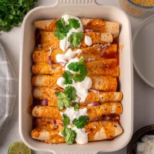 High-protein black bean chicken enchiladas baked in a white casserole dish, topped with melted cheese, sour cream, fresh cilantro, and diced red onion in a rich red enchilada sauce.