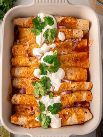 High-protein black bean chicken enchiladas baked in a white casserole dish, topped with melted cheese, sour cream, fresh cilantro, and diced red onion in a rich red enchilada sauce.