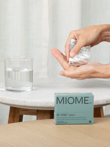 Person holding a Miome IB-ONE probiotic supplement capsule above a glass of water on a small marble table.